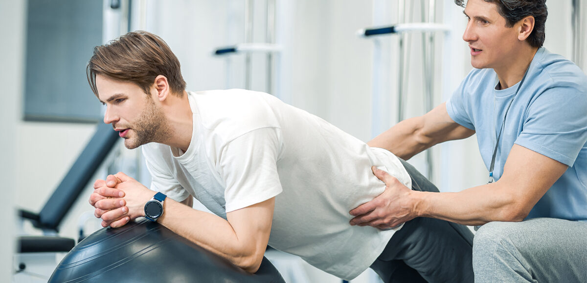 Male physical therapist helps male athletic patient balance on a therapy exercise ball.