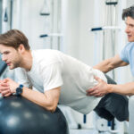 Male physical therapist helps male athletic patient balance on a therapy exercise ball.