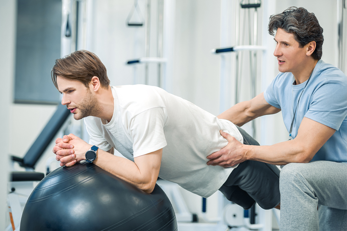 Male physical therapist helps male athletic patient balance on a therapy exercise ball.