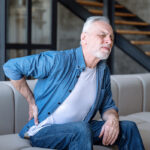 Elder man holding his hip in pain while sitting in his home sofa