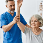 Male physical therapists helps elderly female patient lift her arm while doing manual therapy during a physical assessment