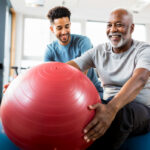A physical therapist works with an elder male black patient, helping him use an exercise ball to improve flexibility, balance, and core strength.