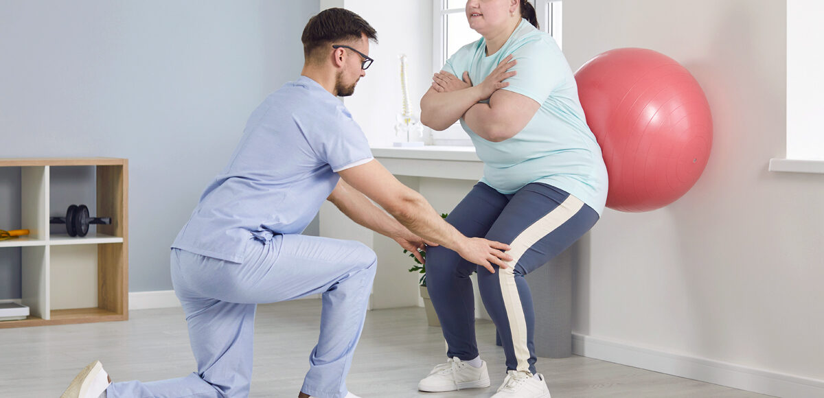 A male physical therapist helps a young overweight woman do wall squats with fit ball.