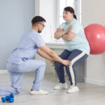 A male physical therapist helps a young overweight woman do wall squats with fit ball.