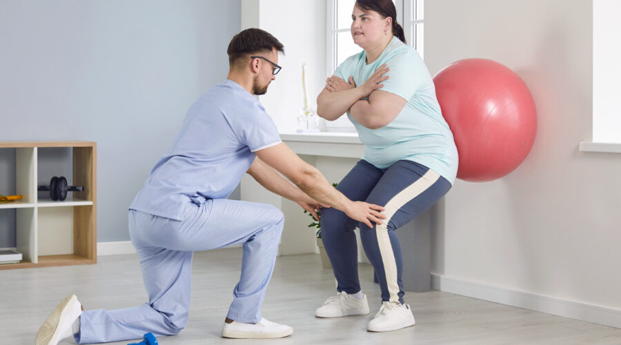 A male physical therapist helps a young overweight woman do wall squats with fit ball.