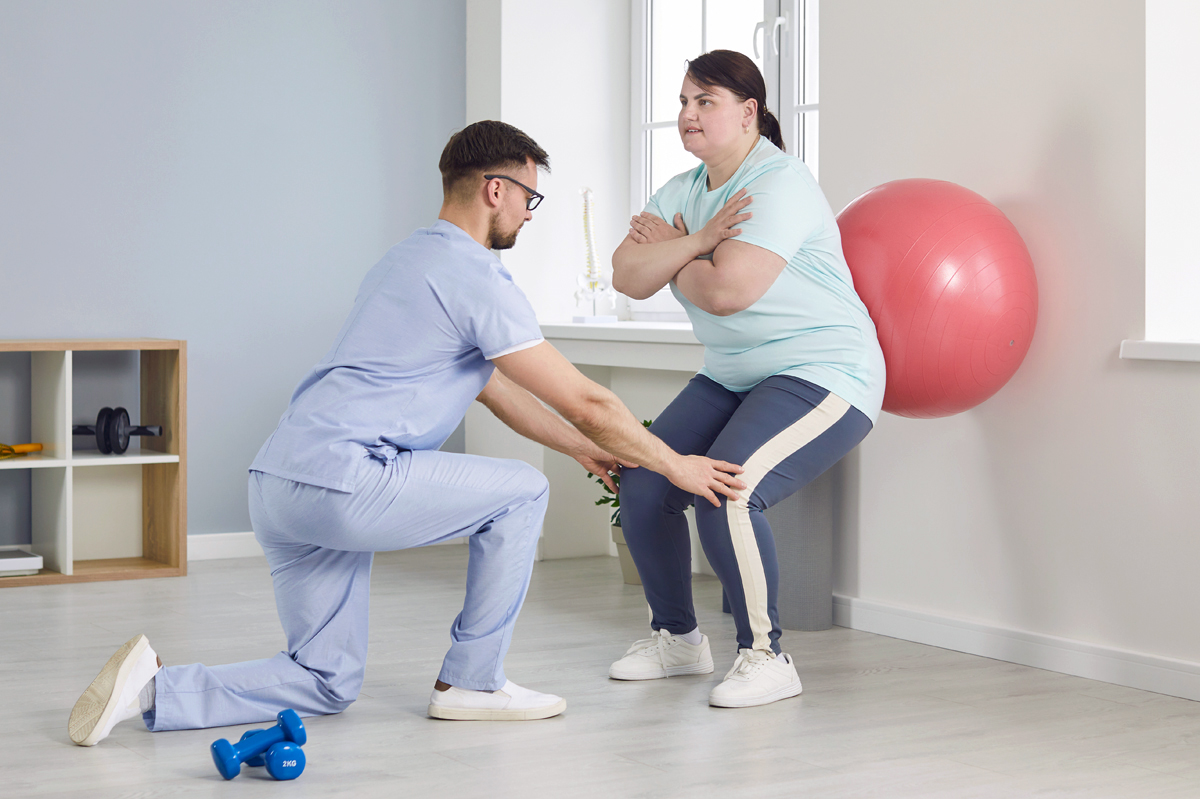 A male physical therapist helps a young overweight woman do wall squats with fit ball.