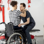 Male physical therapist helping a patient with disabilities standing up from a wheelchair with proper lifting technique as part of their physical therapy rehabilitation program.