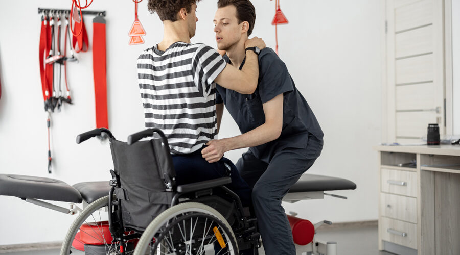 Male physical therapist helping a patient with disabilities standing up from a wheelchair with proper lifting technique as part of their physical therapy rehabilitation program.