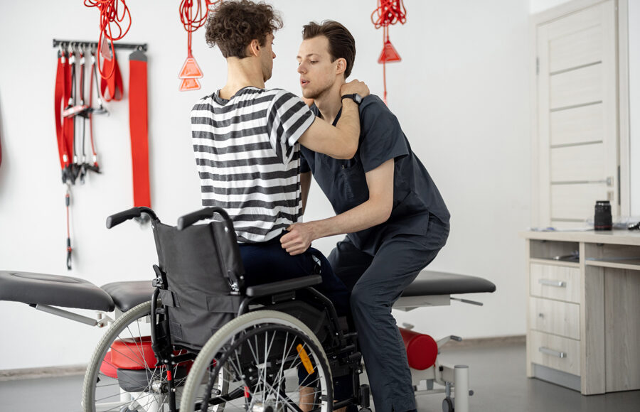 Male physical therapist helping a patient with disabilities standing up from a wheelchair with proper lifting technique as part of their physical therapy rehabilitation program.
