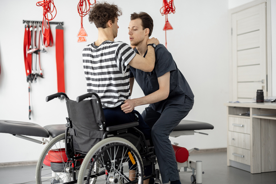 Male physical therapist helping a patient with disabilities standing up from a wheelchair with proper lifting technique as part of their physical therapy rehabilitation program.