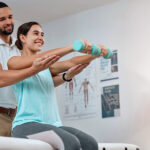 Young woman sitting on a exercise table doing dumbbells with the help of male physical therapist