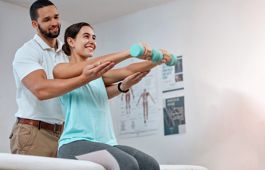 Young woman sitting on a exercise table doing dumbbells with the help of male physical therapist