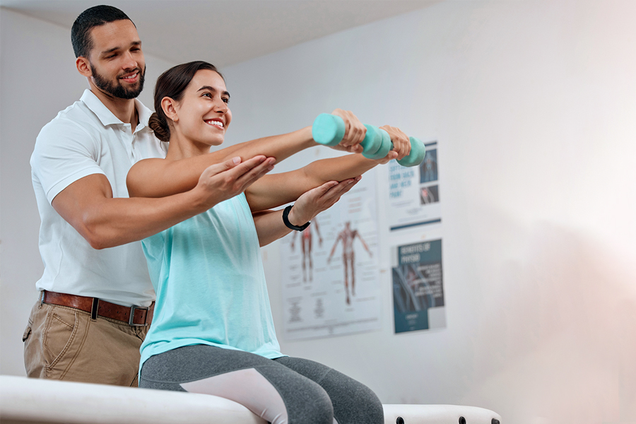 Young woman sitting on a exercise table doing dumbbells with the help of male physical therapist