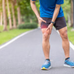 A young adult male stops and holds his knee do to muscle pain while running.
