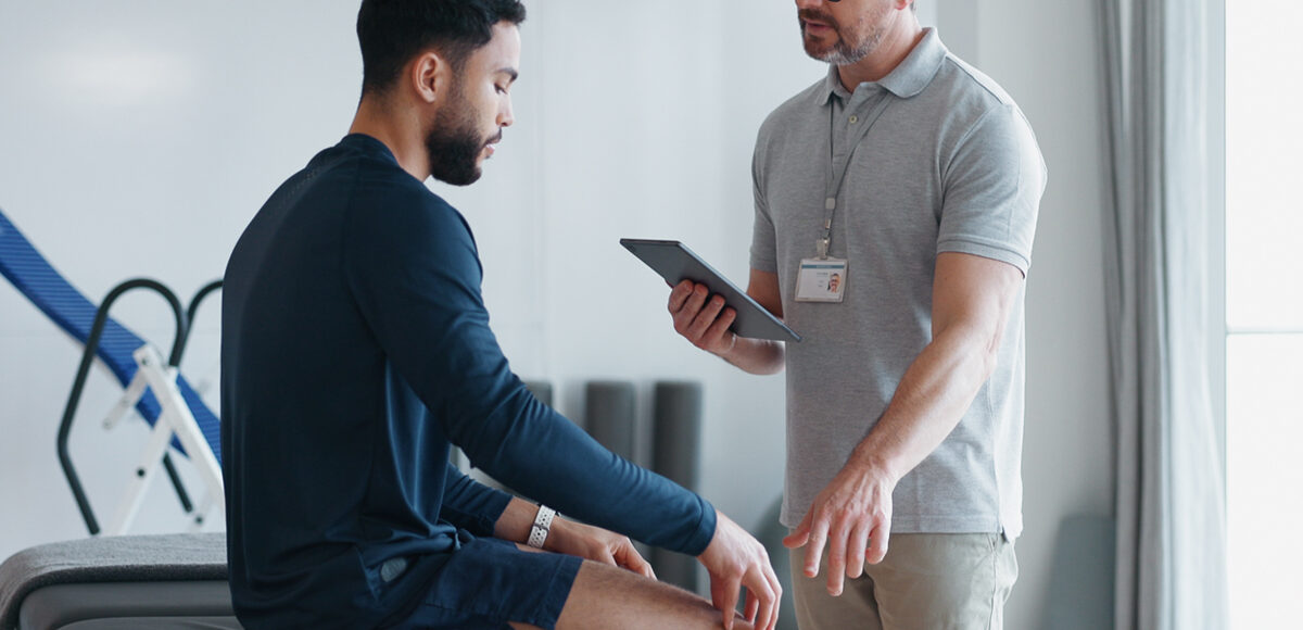 Male physical therapist conducting a physical assessment of a young male patient to evaluate mobility, strength, and range of motion.