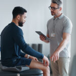 Male physical therapist conducting a physical assessment of a young male patient to evaluate mobility, strength, and range of motion.
