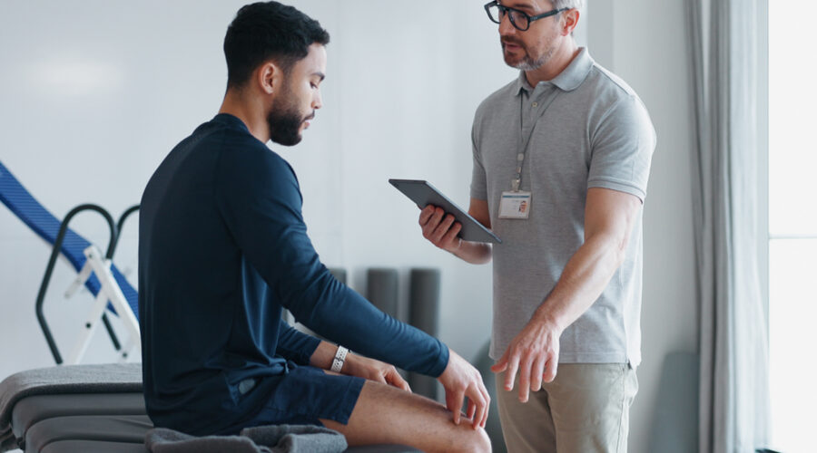 Male physical therapist conducting a physical assessment of a young male patient to evaluate mobility, strength, and range of motion.